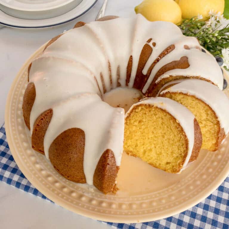 A glazed lemon bundt cake with a slice cut out, displayed on a cream-colored plate with lemons and flowers in the background.