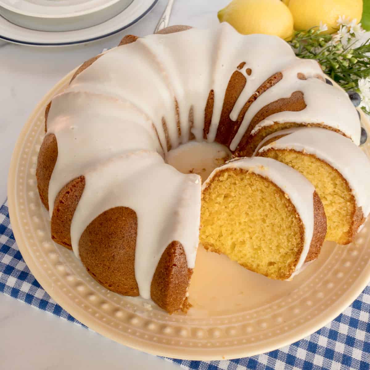 A glazed lemon bundt cake with a slice cut out, displayed on a cream-colored plate with lemons and flowers in the background.