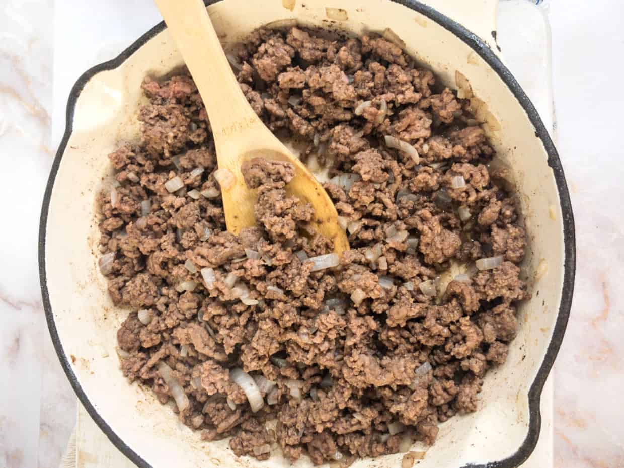 Cooked ground beef and chopped onions in a white skillet being stirred with a wooden spoon.