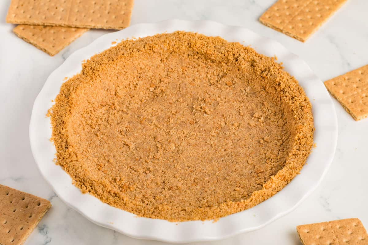 A white pie dish filled with a pressed graham cracker crust, surrounded by several rectangular graham crackers on a light surface.