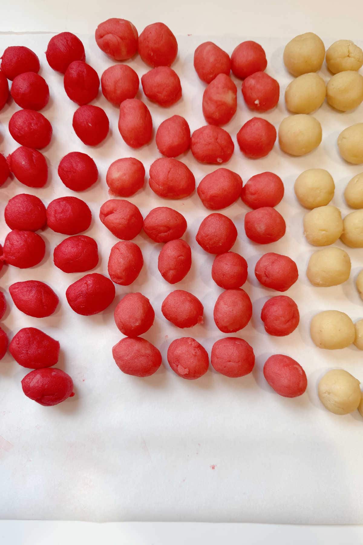 Rows of small red and beige cookie dough balls are arranged on a sheet of white parchment paper.