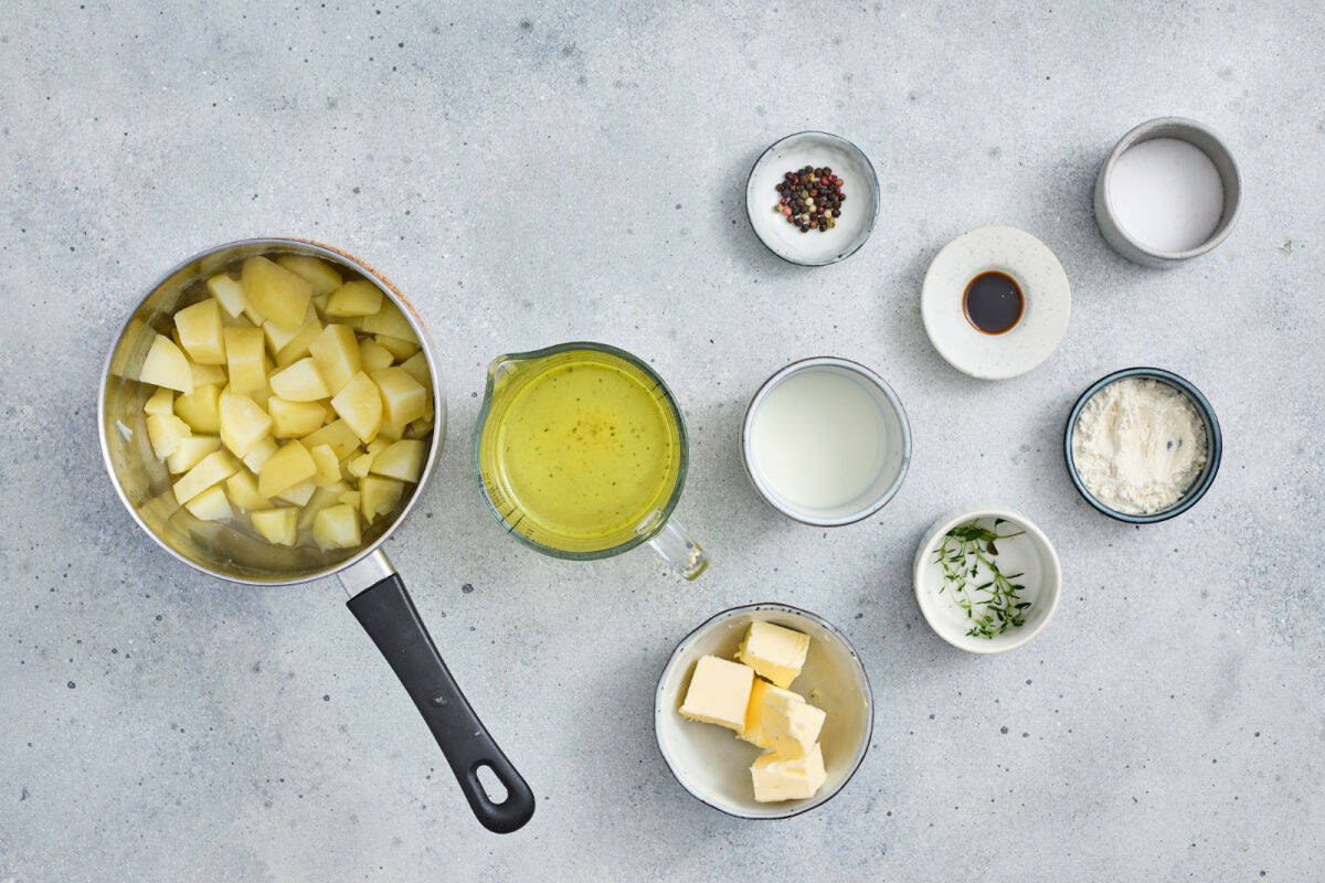 Overhead view of prepped ingredients for mashed potatoes, including chopped potatoes in a pot, butter, milk, cream, broth, salt, pepper, herbs, and Worcestershire sauce on a gray surface.