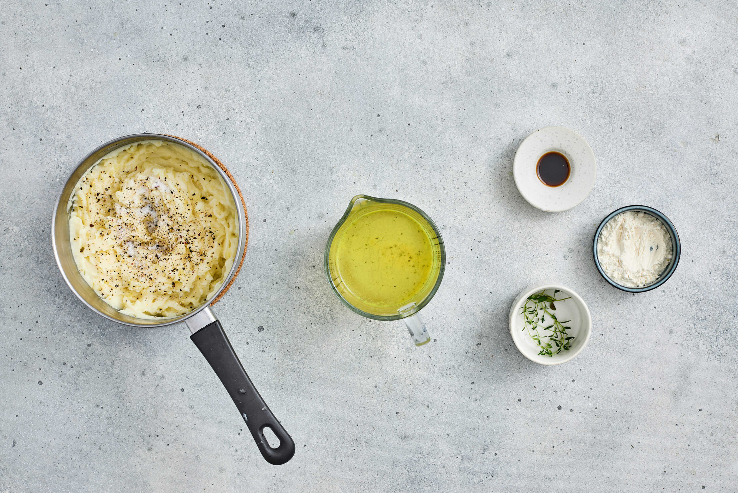 A saucepan with mashed potatoes, a measuring cup of yellow liquid, and small bowls of soy sauce, herbs, and grated cheese on a light gray surface.