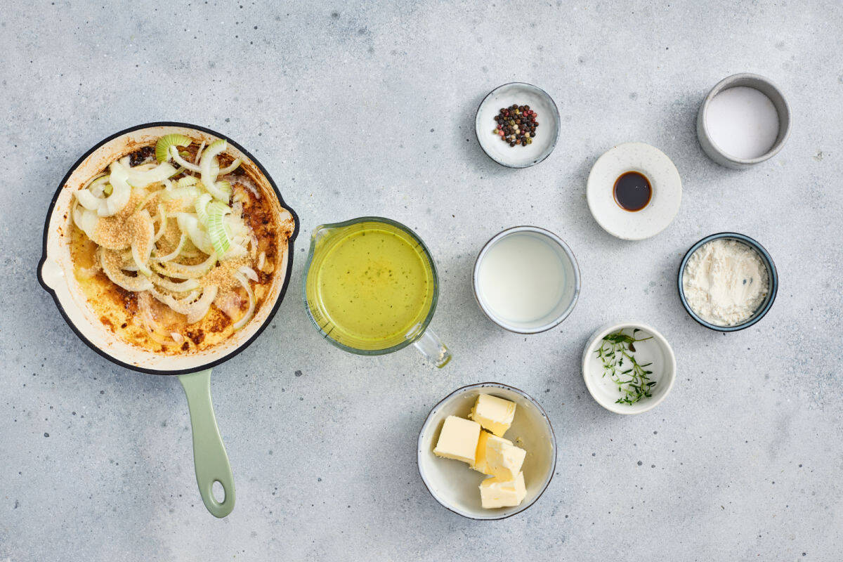 A skillet with cooked onions sits beside bowls of butter, flour, milk, broth, fresh herbs, soy sauce, salt, and pepper on a light gray surface.