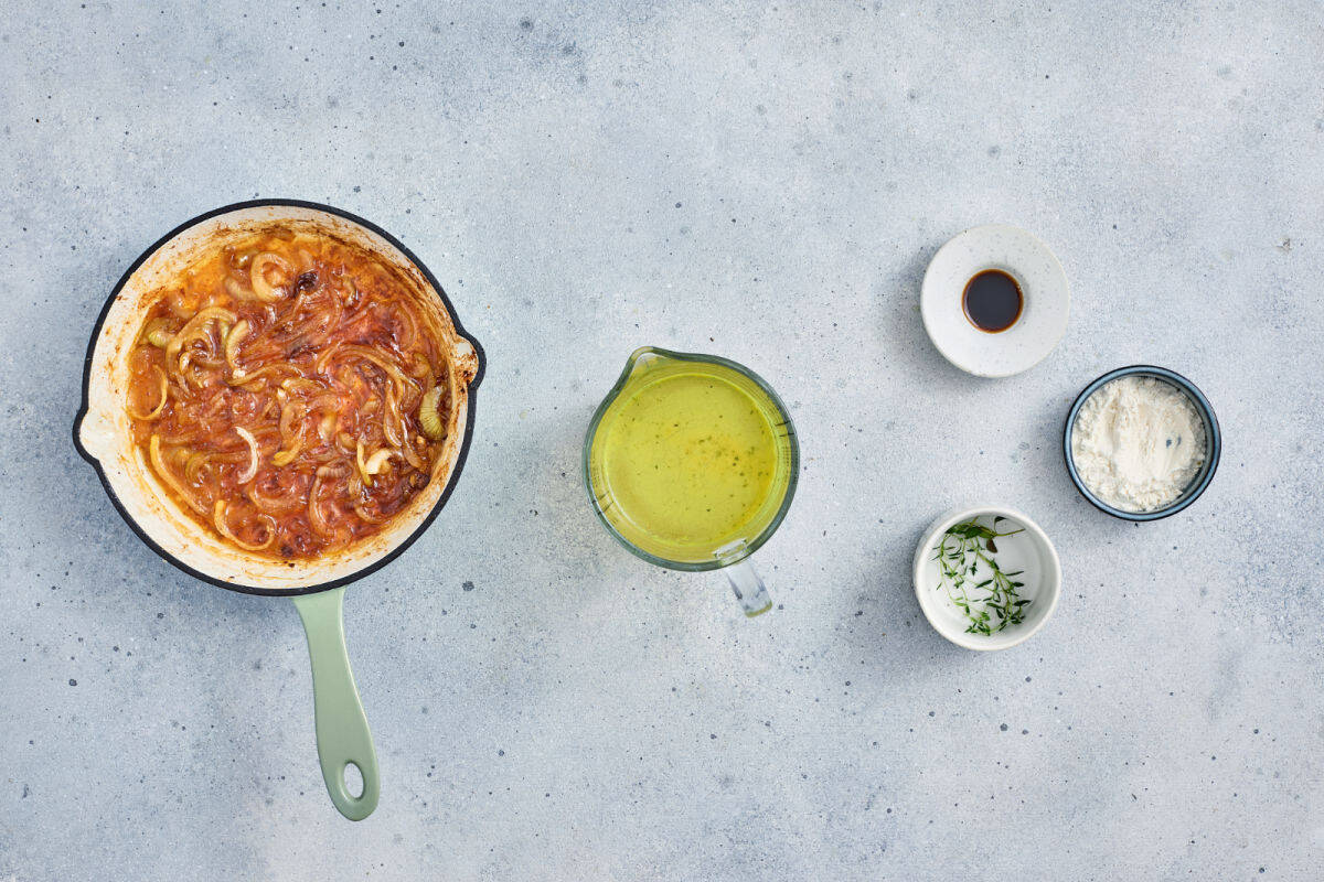 A pan with cooked onions and tomato sauce, a jug of yellow liquid, a small bowl of dark sauce, a bowl of flour, and a bowl of fresh herbs are arranged on a light grey surface.