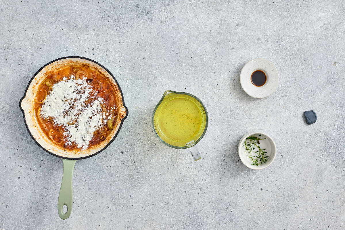 A skillet with onionb and flour, a measuring cup of broth, a small bowl of herbs, and a small dish of dark liquid are arranged on a gray countertop.