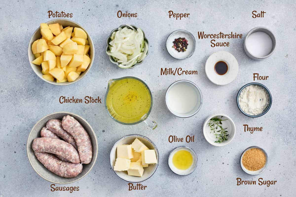 Top-down view of bowls containing raw ingredients for cooking: potatoes, onions, pepper, Worcestershire sauce, salt, chicken stock, milk/cream, flour, thyme, brown sugar, olive oil, butter, and sausages.
