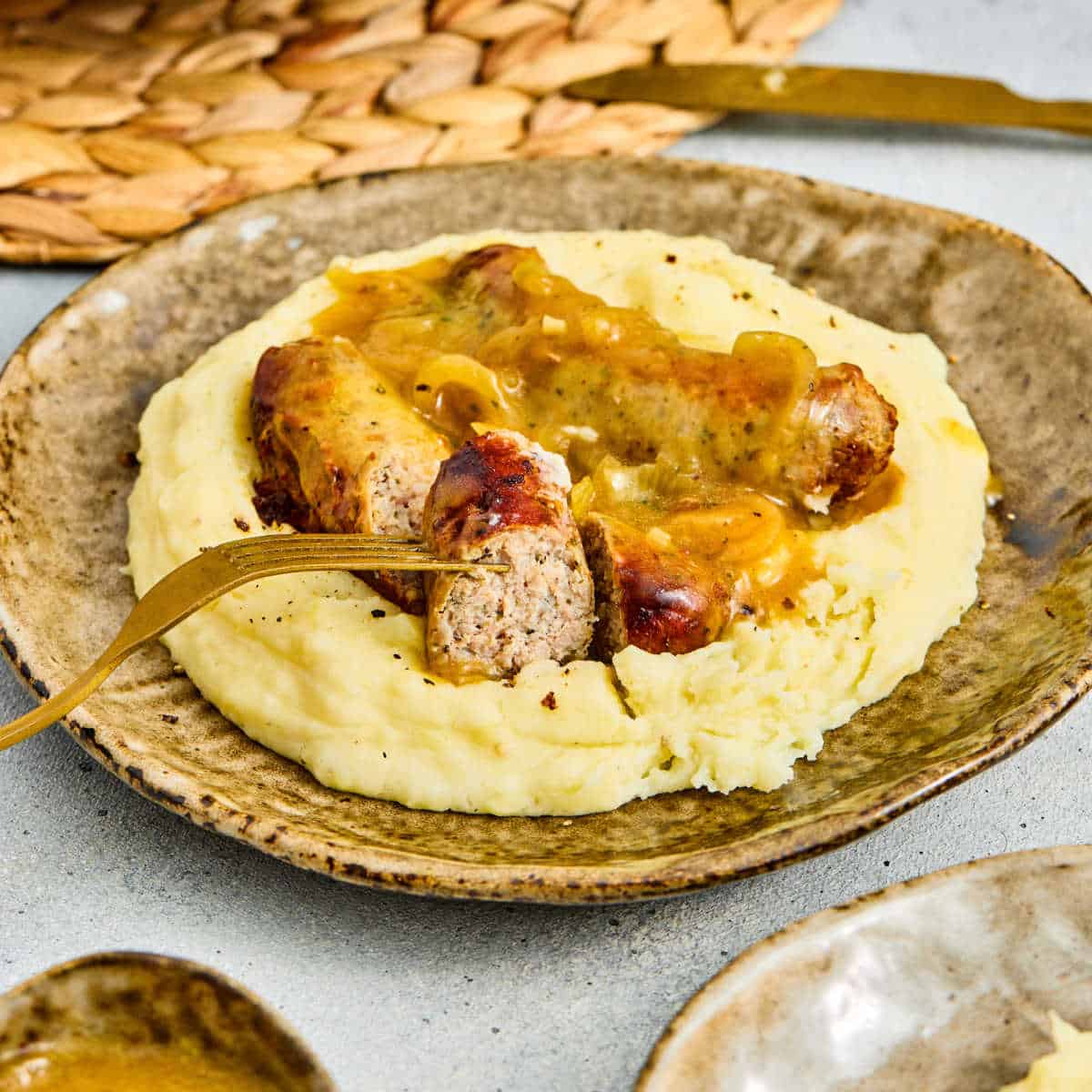 A plate of mashed potatoes topped with sausages and brown onion gravy, with a fork cutting into one sausage.