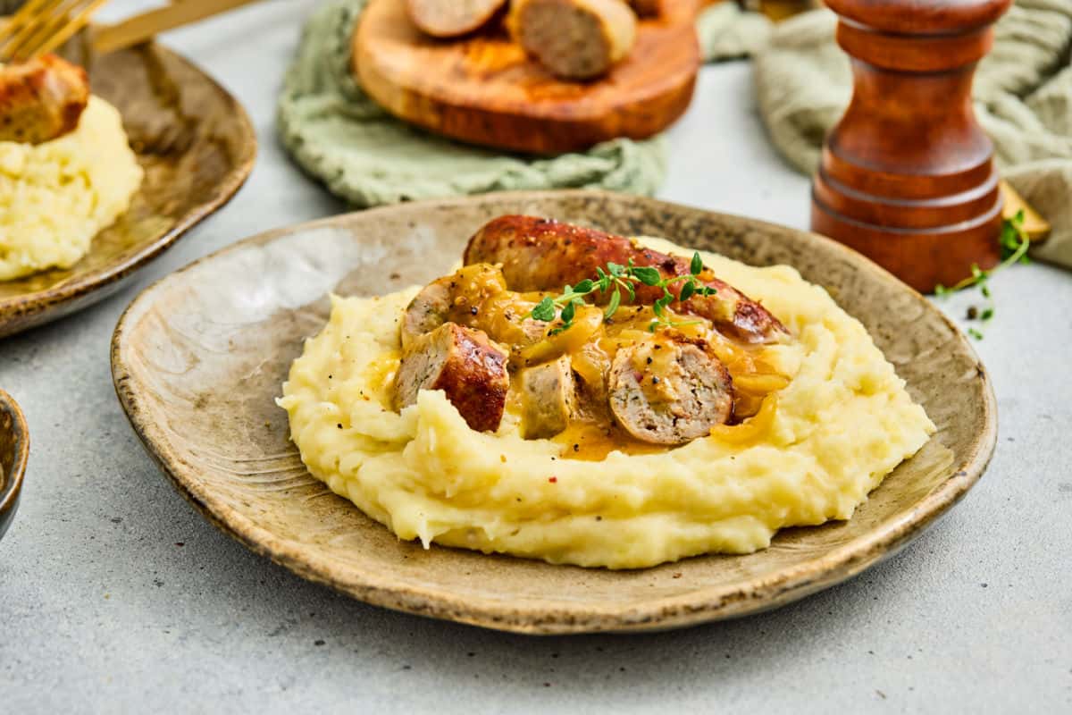 A plate of mashed potatoes topped with sliced sausage and onion gravy, garnished with a sprig of thyme.