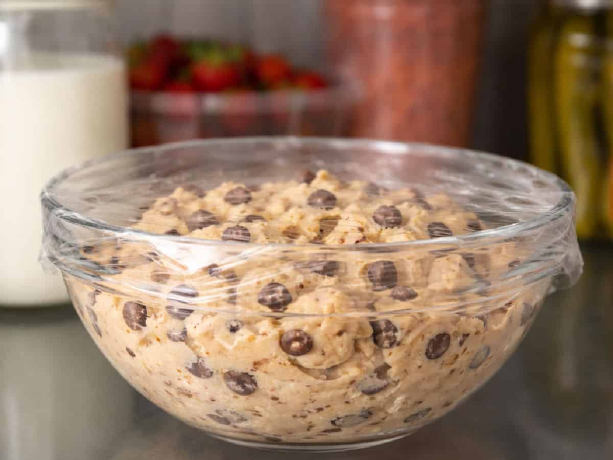 A glass bowl filled with chocolate chip cookie dough is covered with plastic wrap and sits on a counter.