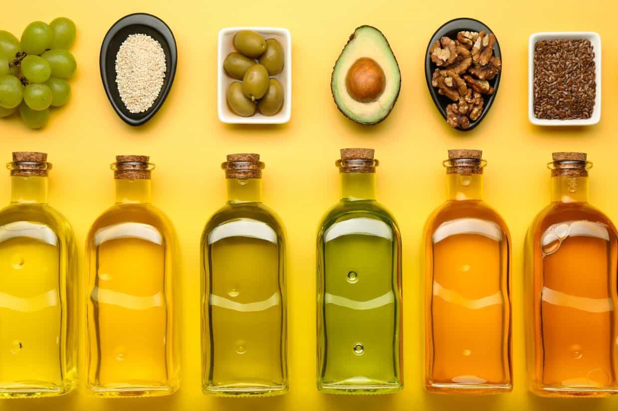 Five bottles of oil are lined up on a yellow background, with ingredients such as grapes, sesame seeds, olives, avocado, walnuts, and flaxseeds arranged in bowls above them.