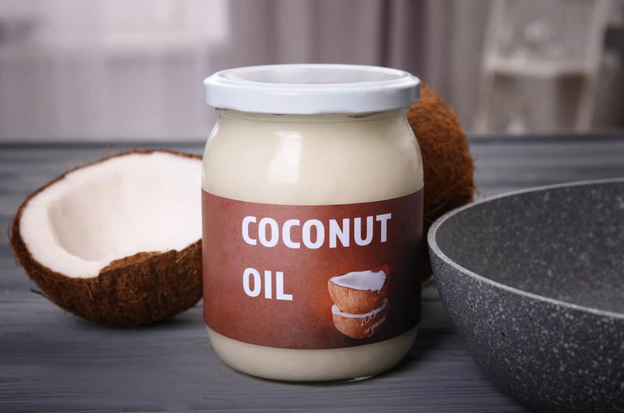 A jar labeled "Coconut Oil" sits on a table next to a halved coconut and a gray pan.