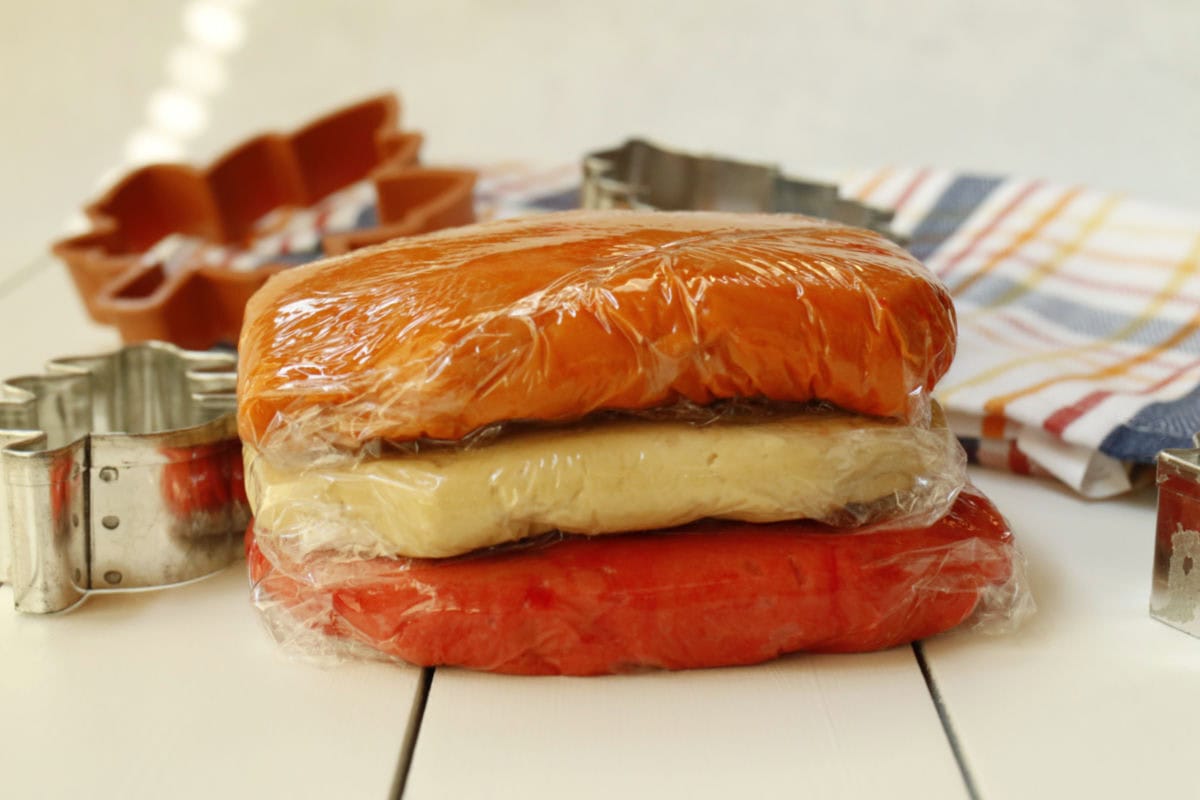 Three stacks of colored cookie dough wrapped in plastic wrap, with metal cookie cutters and a striped cloth in the background on a white surface.
