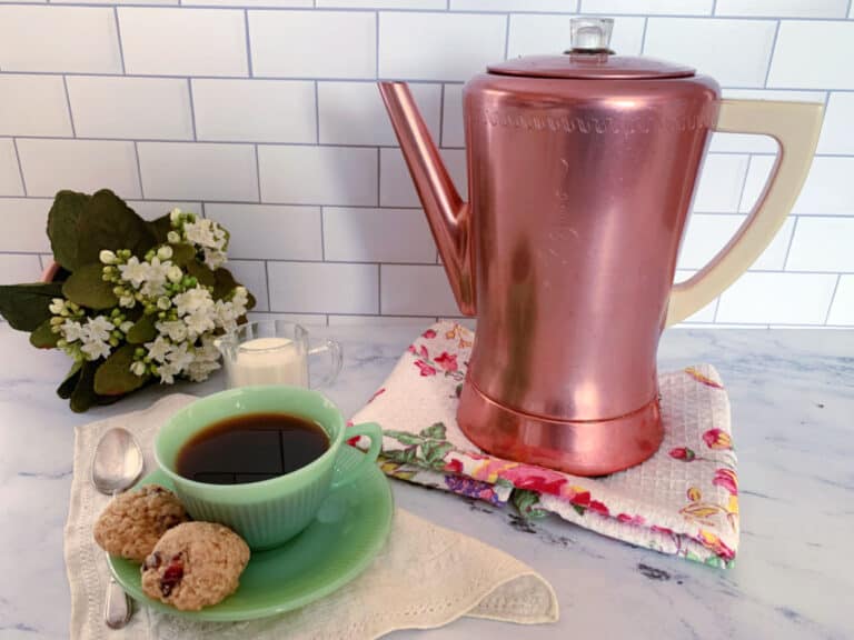 A copper coffee percolator, a green cup of black coffee, two cookies, a spoon, a small glass of milk, and a bouquet of white flowers on a kitchen counter with a tiled backsplash.