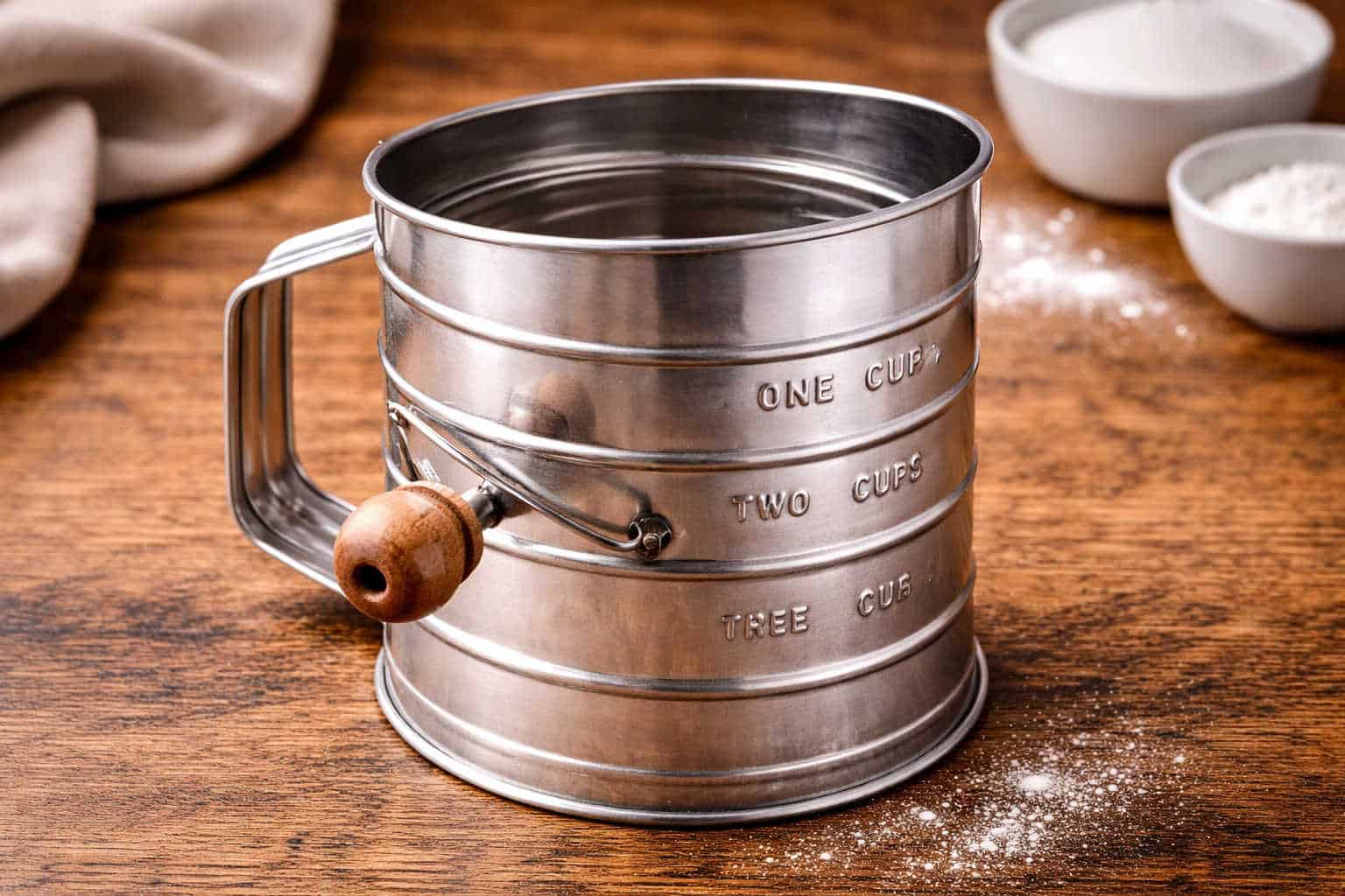 A metal flour sifter with a wooden handle sits on a wooden surface, with measuring marks visible and bowls of flour in the background.