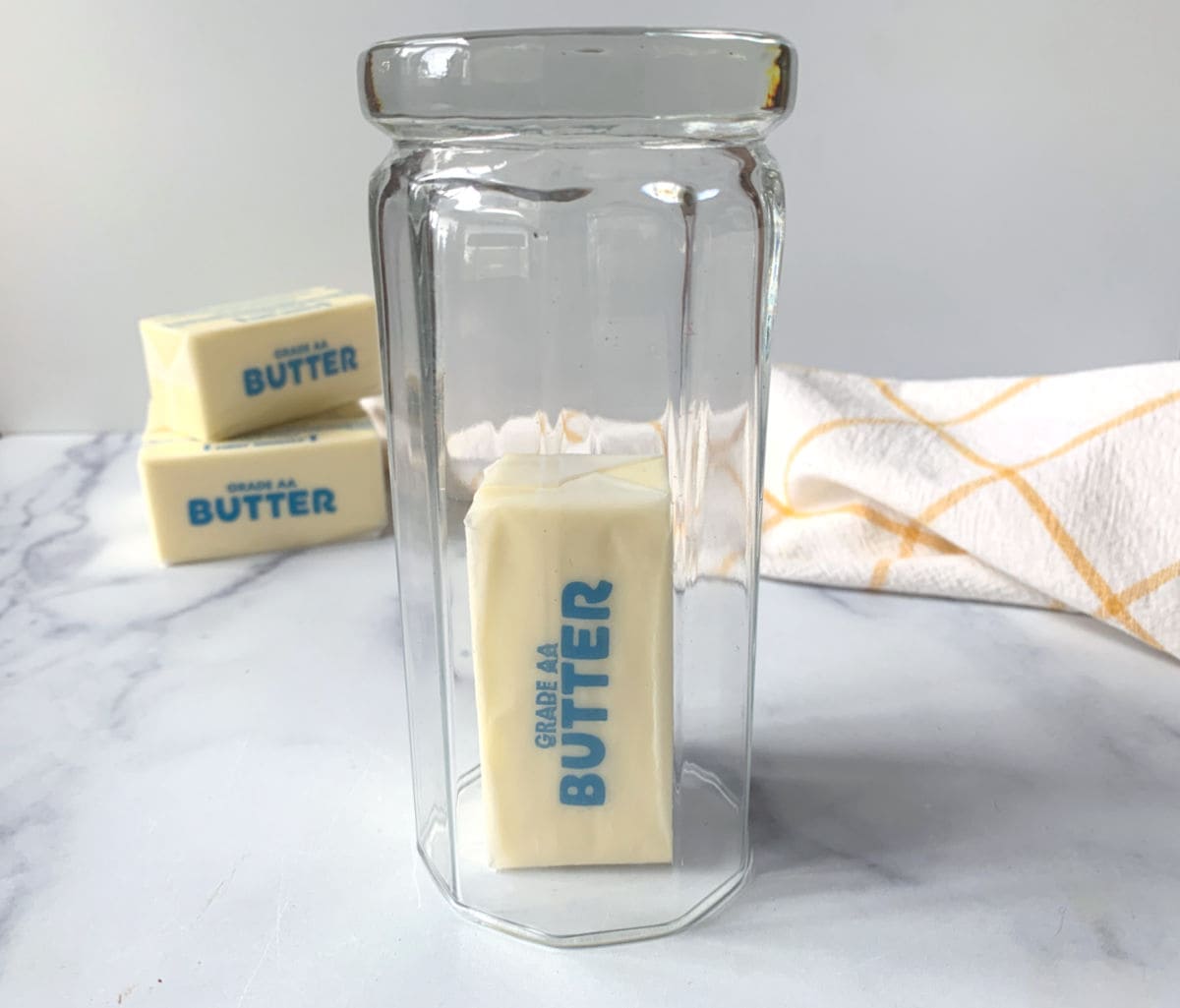 A stick of butter labeled "Grade AA Butter" is standing upright inside a clear glass jar, with two more butter sticks and a yellow-checked towel in the background.