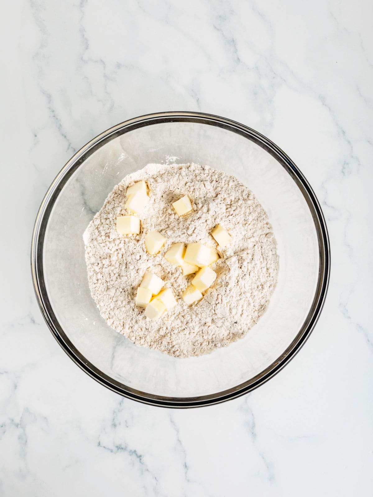 A glass bowl containing flour mixture and cubes of butter sits on a white marble surface.