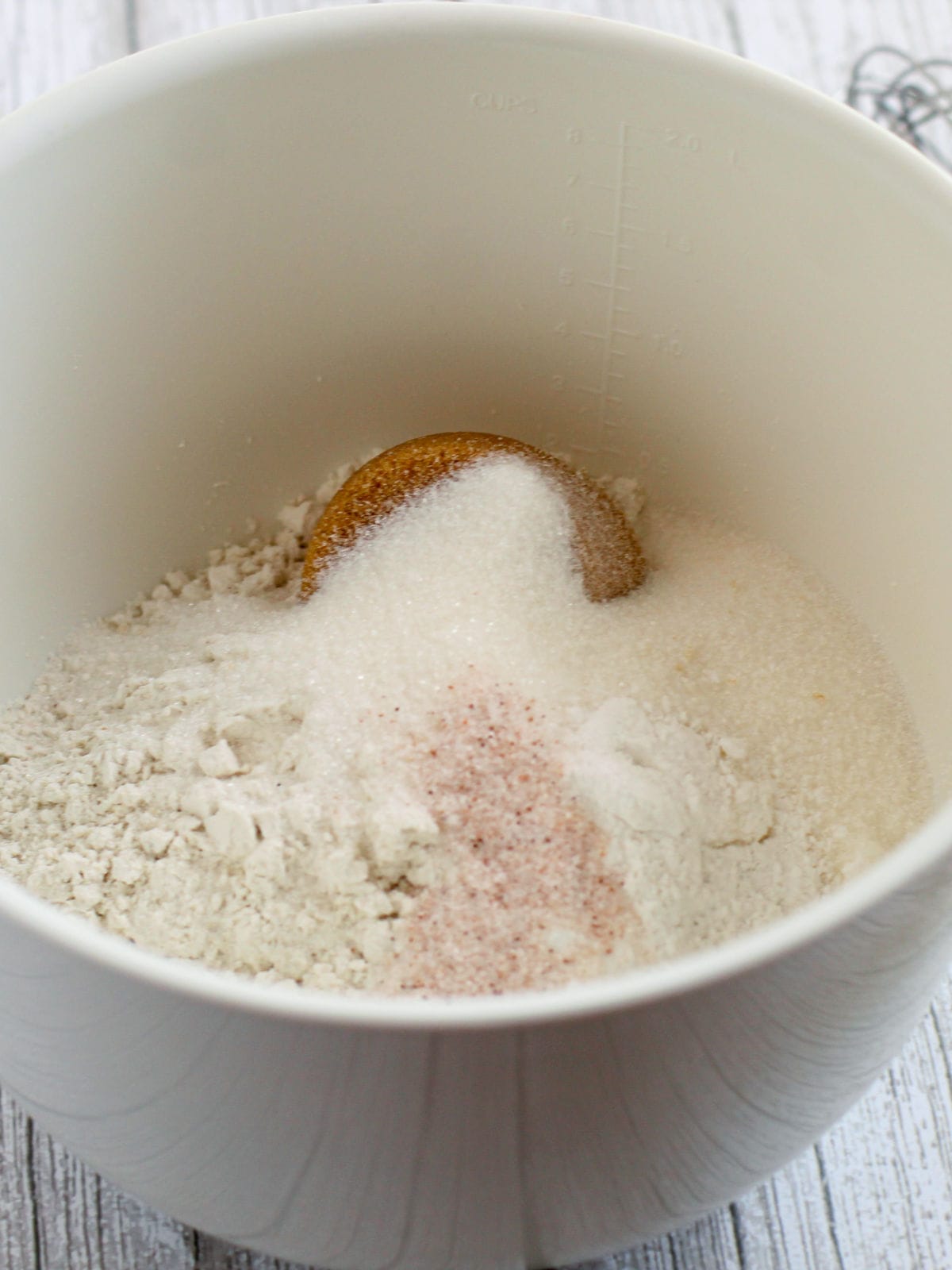 A white mixing bowl containing flour, granulated sugar, brown sugar, and pink salt on a light wooden surface.