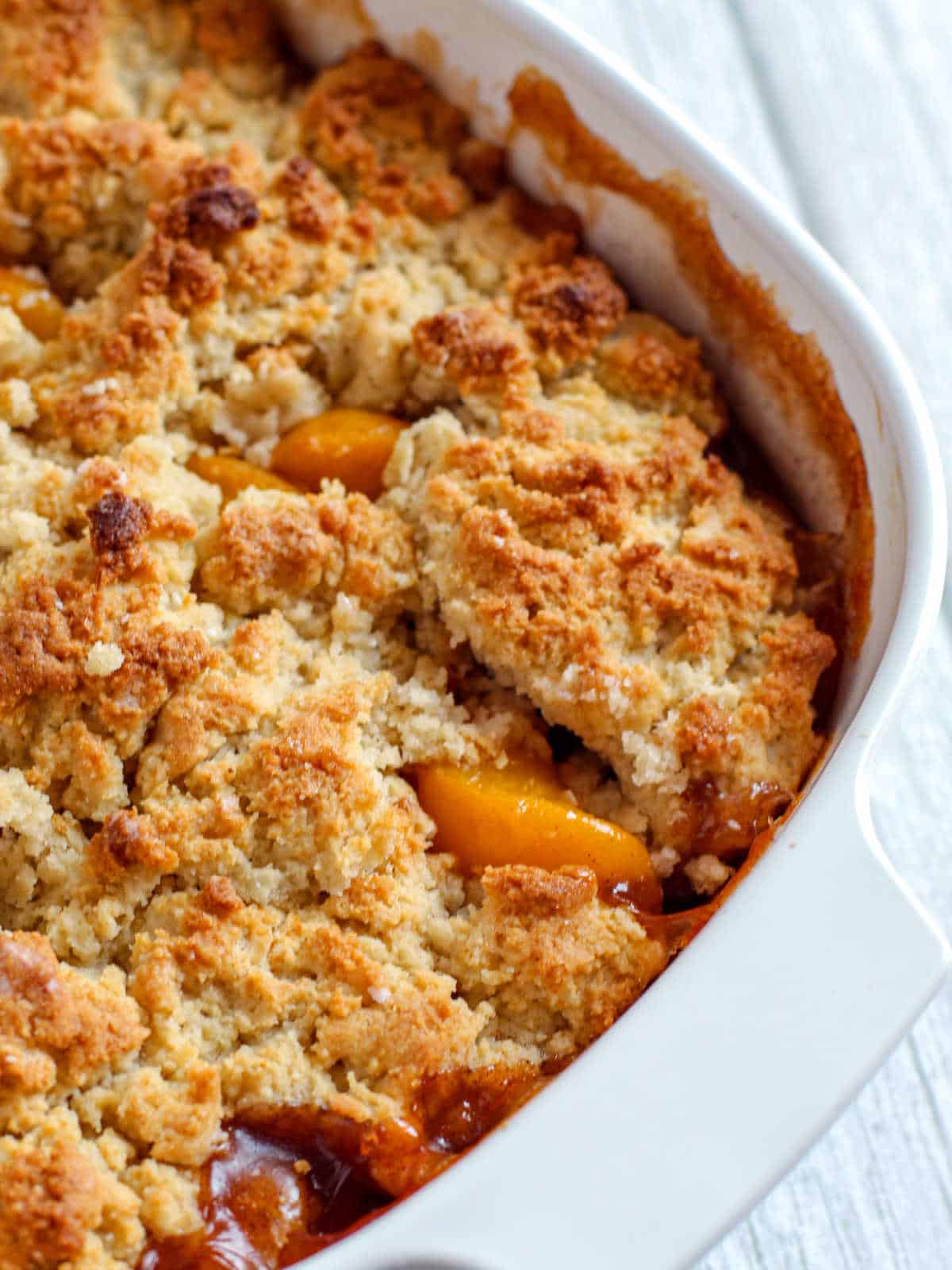 Golden-brown peach cobbler with a crumbly topping in a white baking dish on a light wood surface.