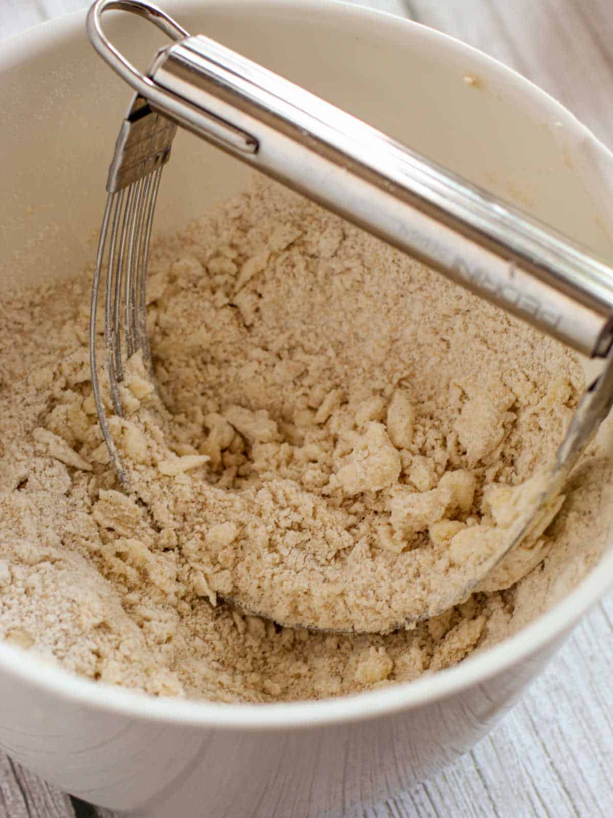 A pastry blender mixing flour and butter in a white bowl to make a crumbly dough mixture.