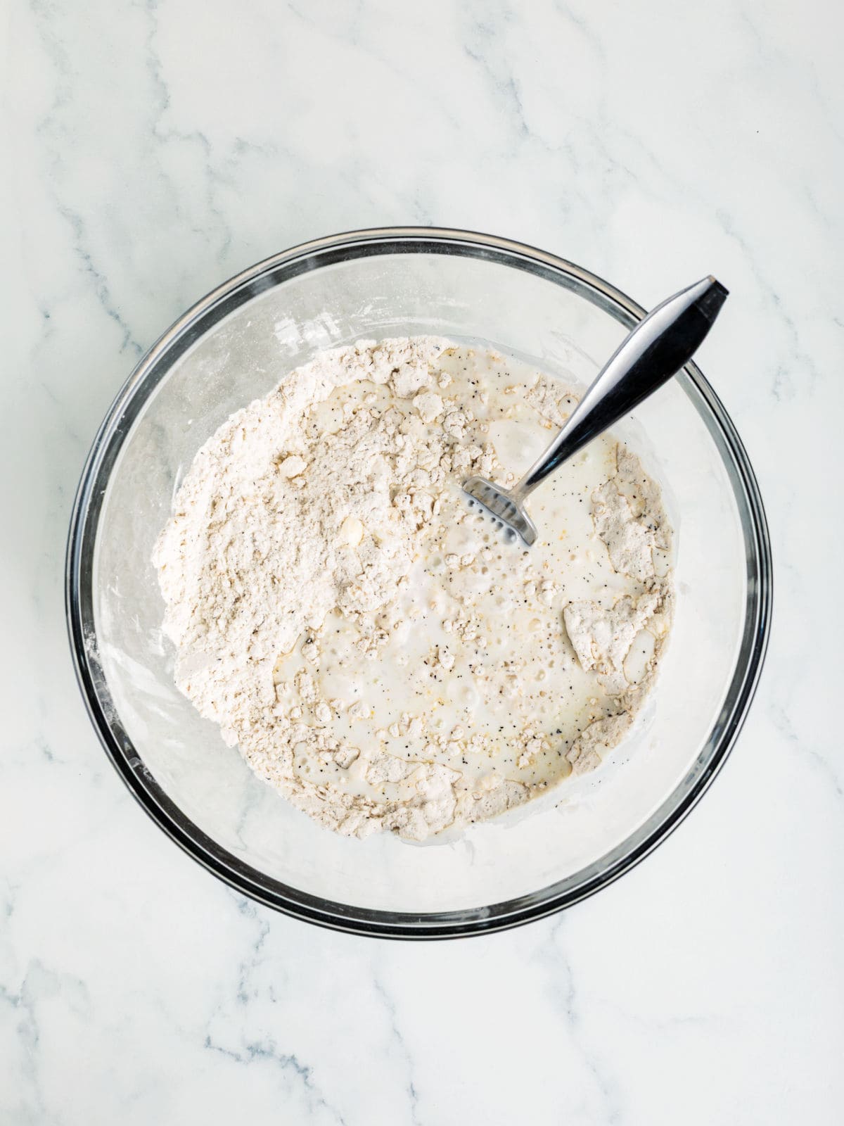 A glass bowl containing flour, pepper, and liquid being mixed with a metal fork on a white marble surface.