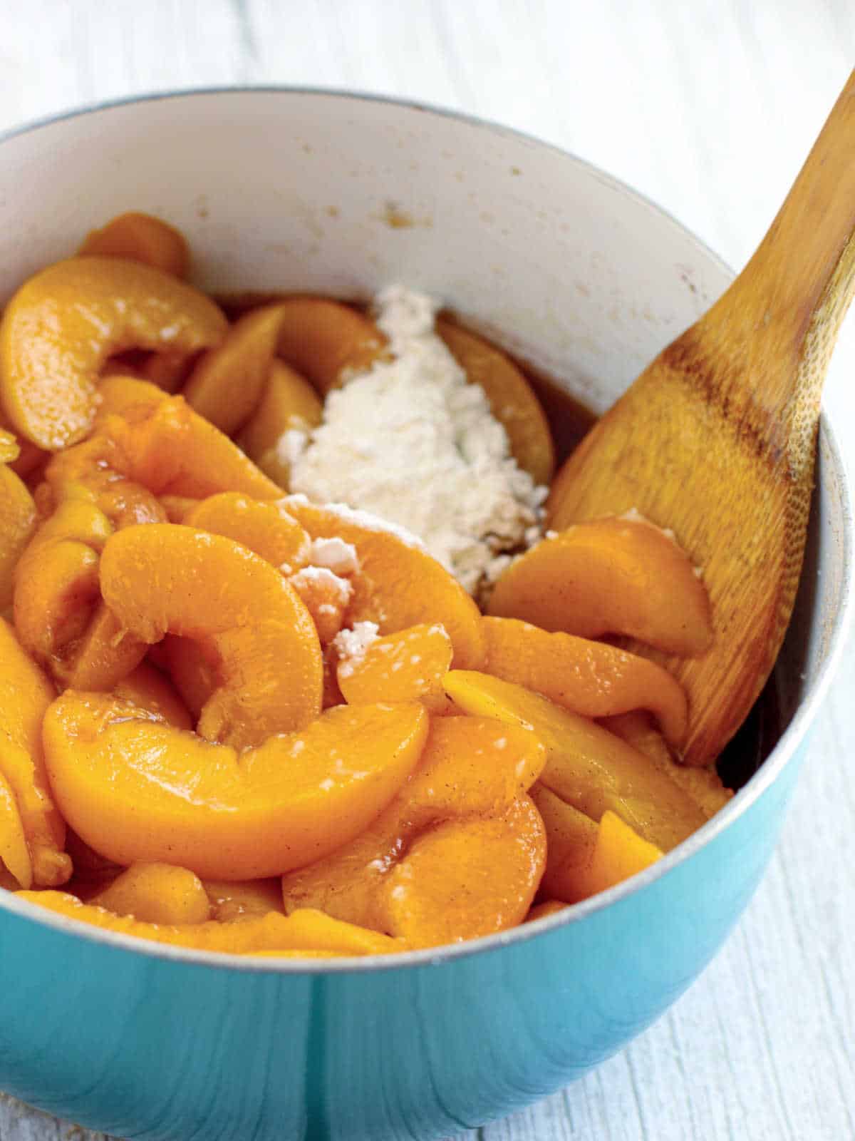 Sliced peaches and a mound of flour are mixed together in a blue pot with a wooden spoon on a light surface.