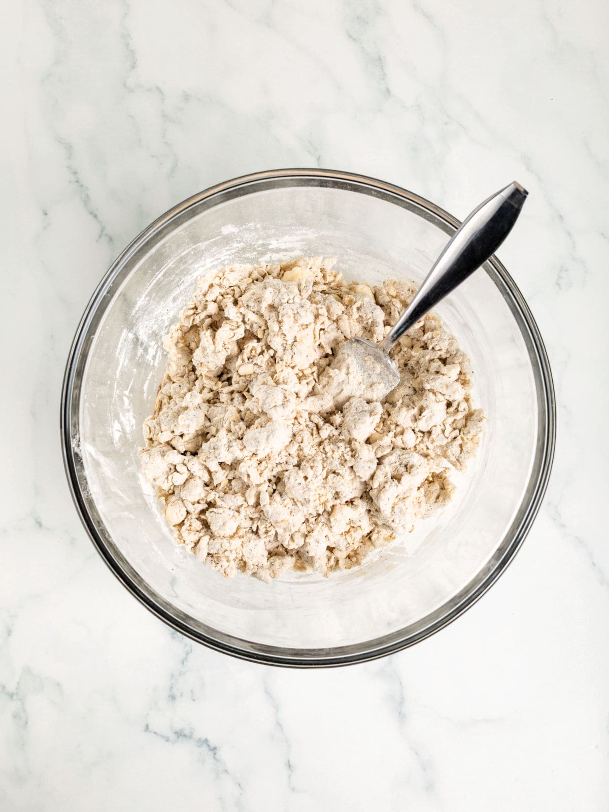 A glass bowl containing partially mixed biscuit dough with a spoon resting inside, on a white marble surface.