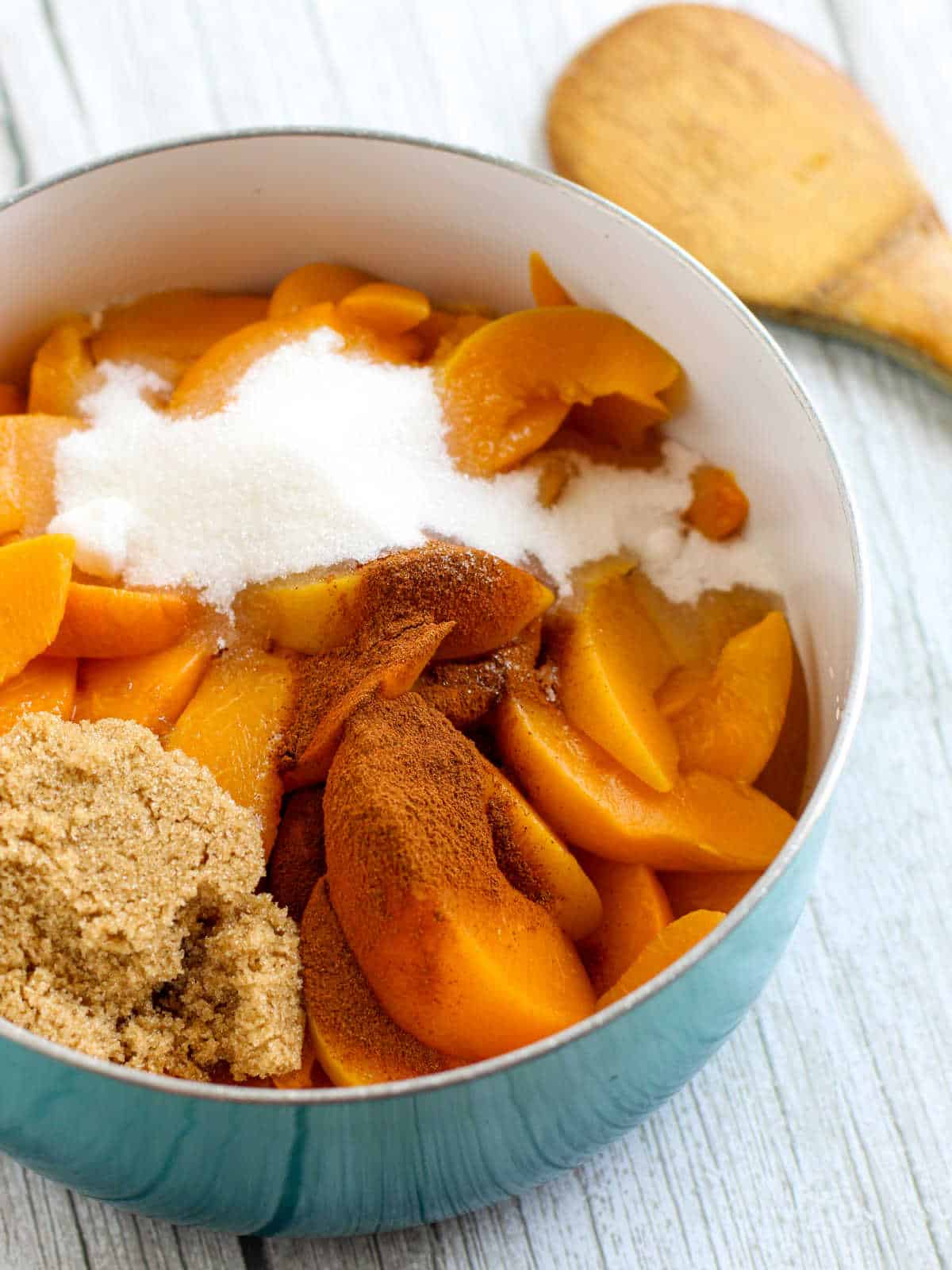 A bowl of sliced peaches topped with granulated sugar, brown sugar, and ground cinnamon, with a wooden spoon nearby on a light wooden surface.