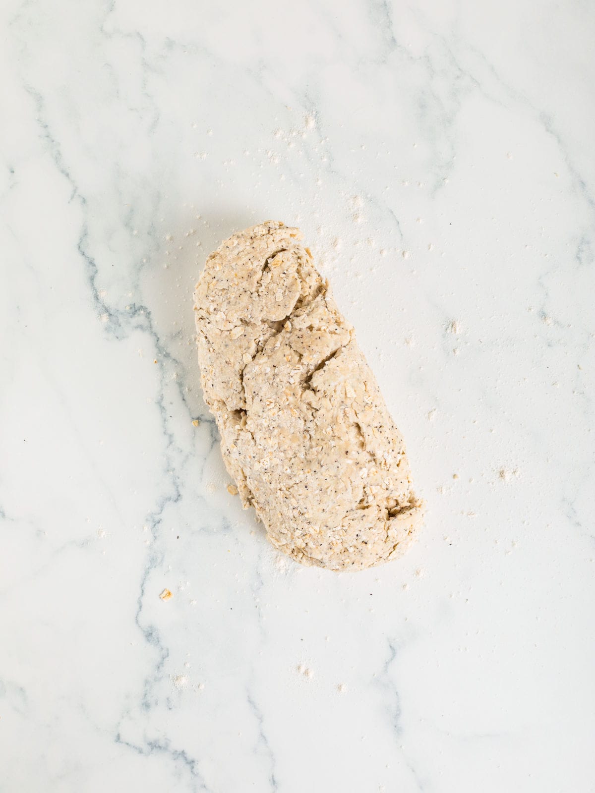 A portion of rough, unshaped dough rests on a white marble surface, with scattered flour and crumbs around it.