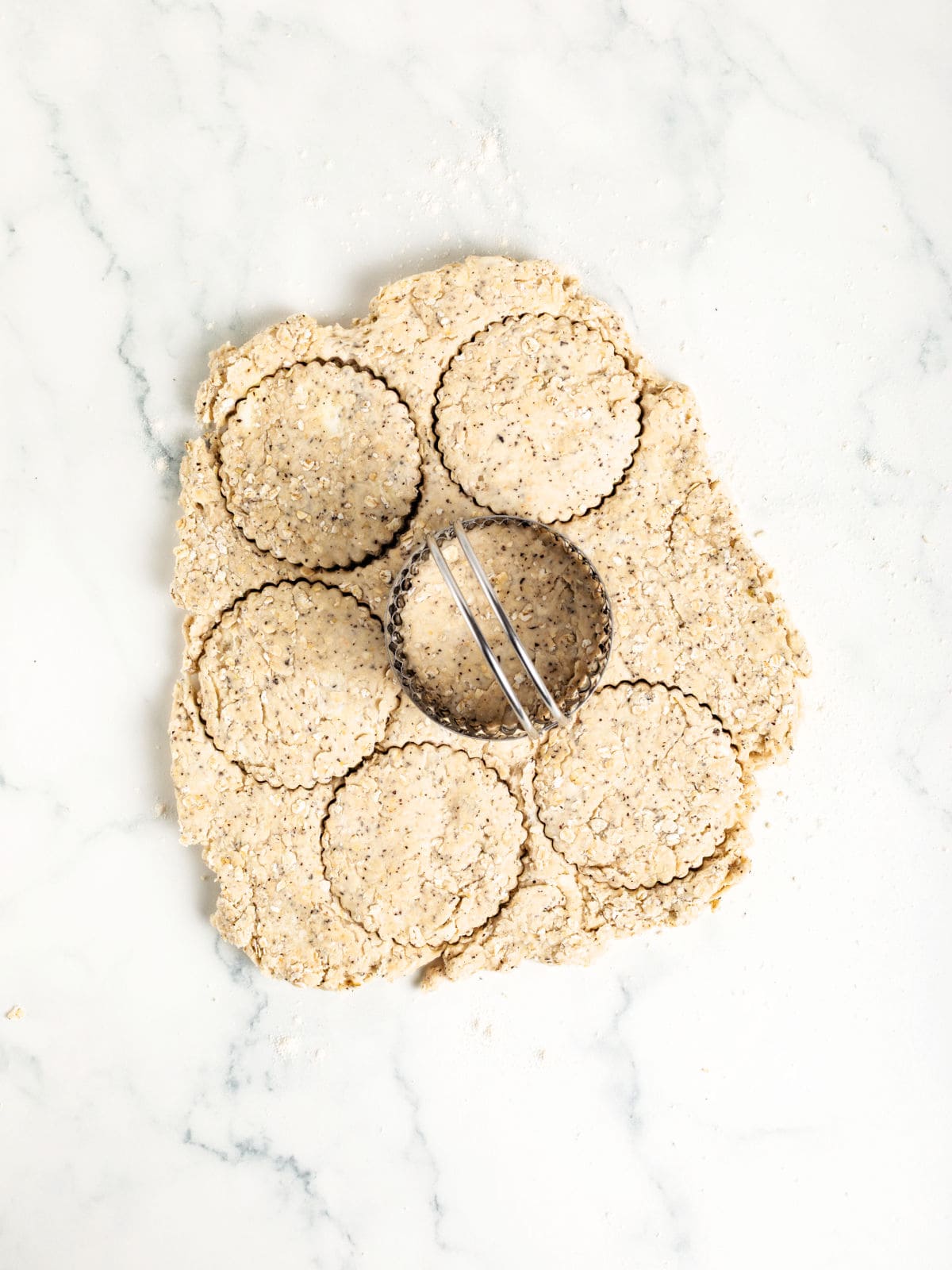A piece of dough with poppy seeds is on a marble surface. Six round shapes have been cut out with a fluted biscuit cutter, which rests on the dough.
