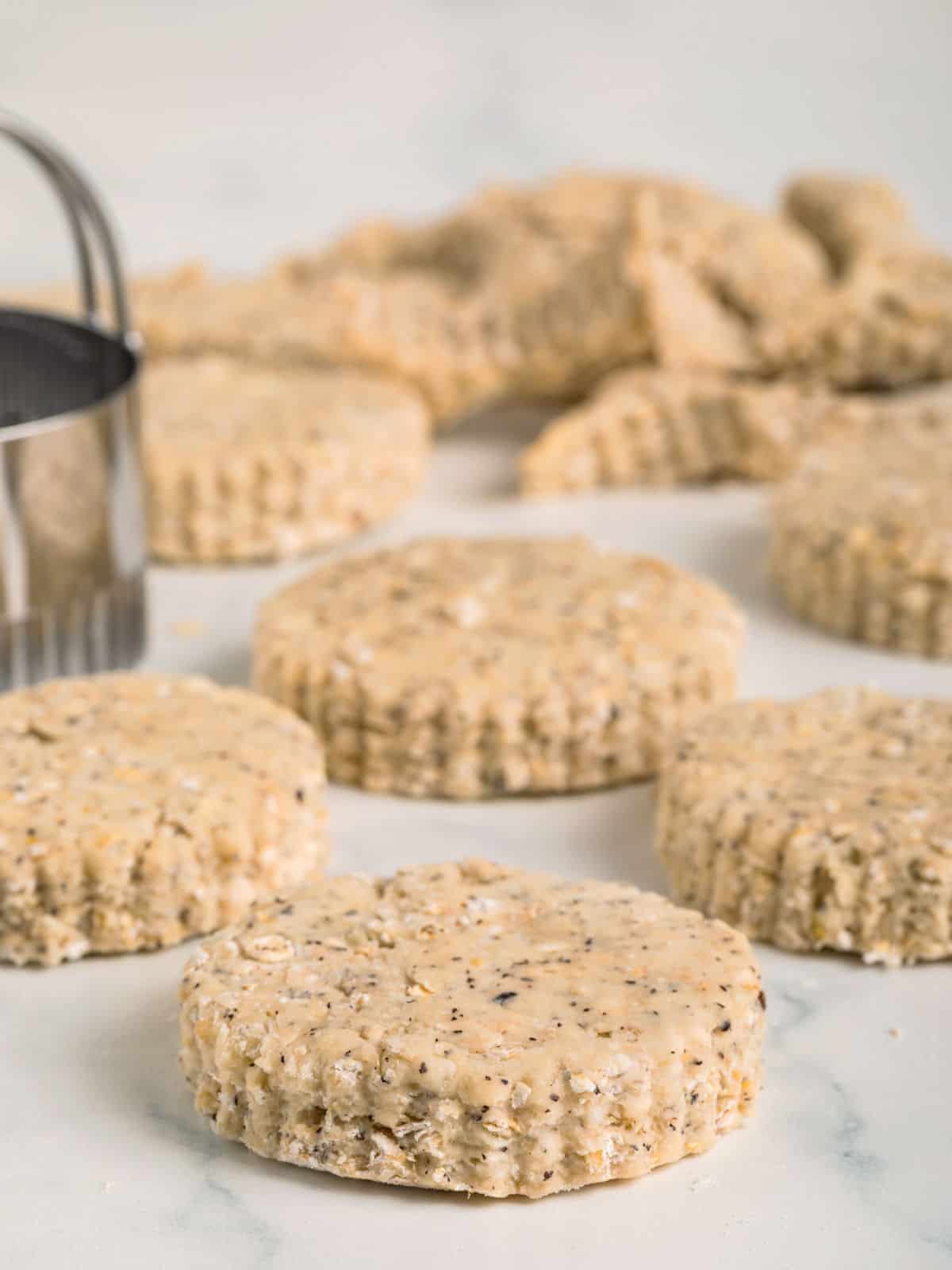 Unbaked round biscuit dough with visible seeds and grains on a floured surface, with a metal biscuit cutter nearby and more dough in the background.