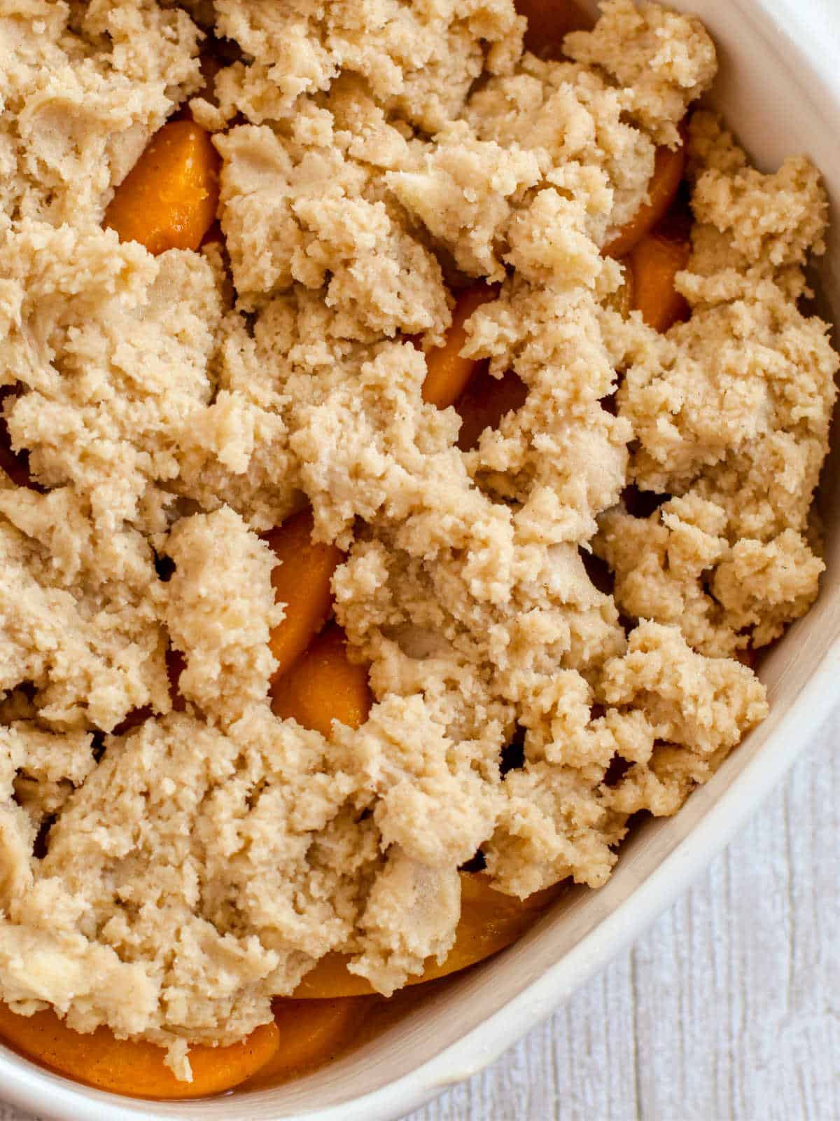 Close-up of an unbaked peach cobbler in a white dish, showing sliced peaches partially covered by a crumbly dough topping.