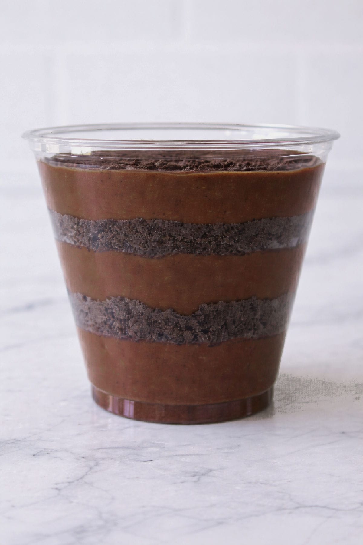 A clear plastic cup filled with alternating layers of chocolate pudding and crushed chocolate cookies on a marble surface.
