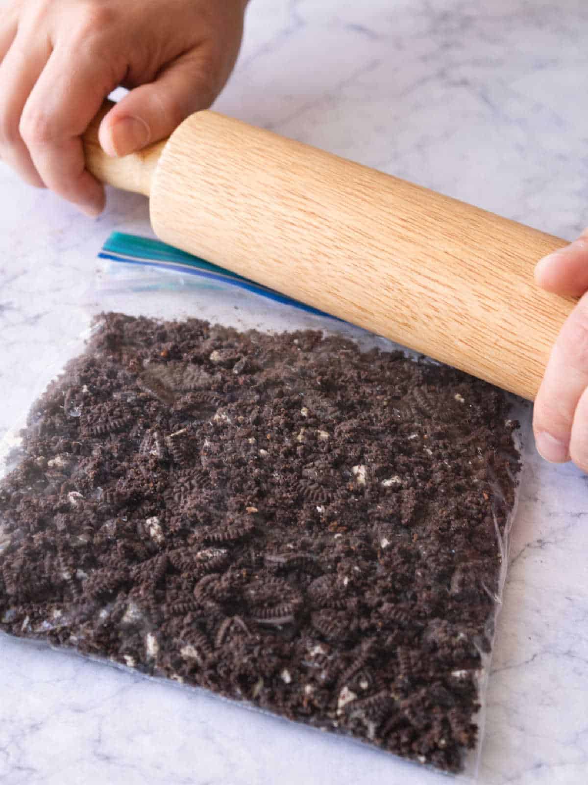 Hands use a rolling pin to crush chocolate cookies inside a sealed plastic bag on a marble countertop.