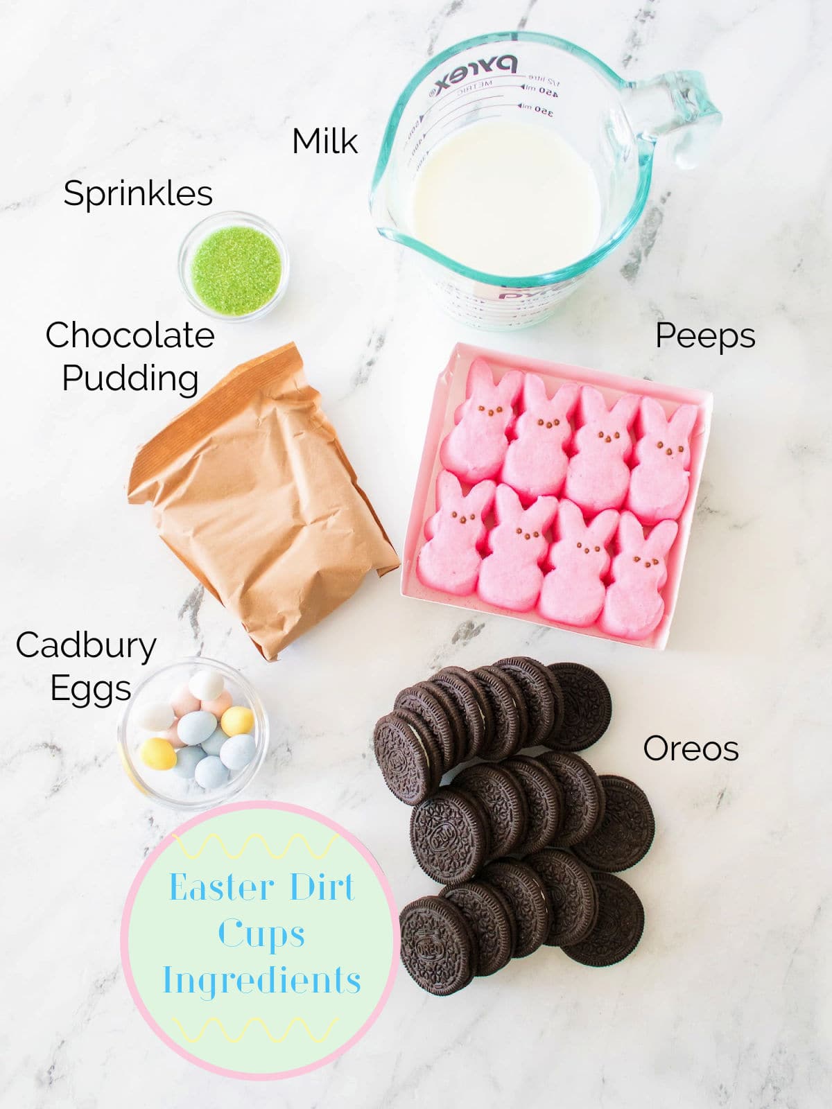 Top-down view of ingredients for Easter dirt cups: milk, green sprinkles, chocolate pudding mix, pink Peeps, Cadbury eggs, and Oreo cookies arranged on a marble surface.