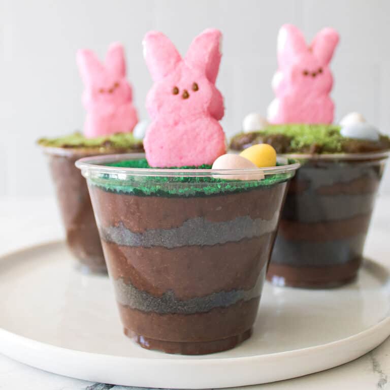 Plastic cups filled with layered chocolate pudding, topped with candy eggs, green sprinkles, and pink marshmallow bunny Peeps, arranged on a white plate.