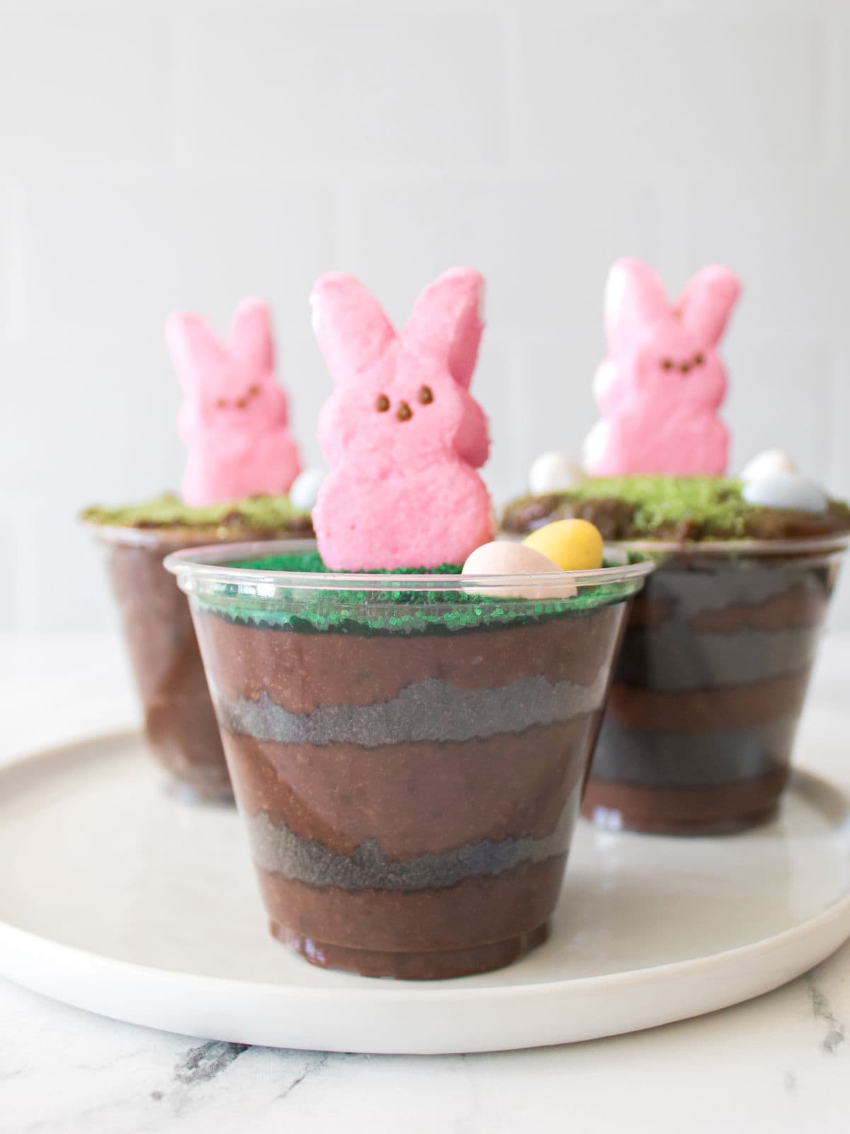 Three clear cups filled with layered chocolate pudding and cookie crumbs, topped with pink marshmallow bunny candies and small candy eggs, are arranged on a white plate.