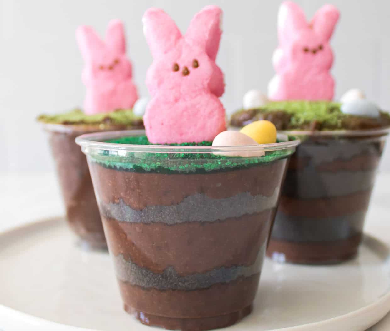 Clear plastic cups filled with layered chocolate pudding and topped with green sprinkles, candy eggs, and a pink marshmallow bunny.