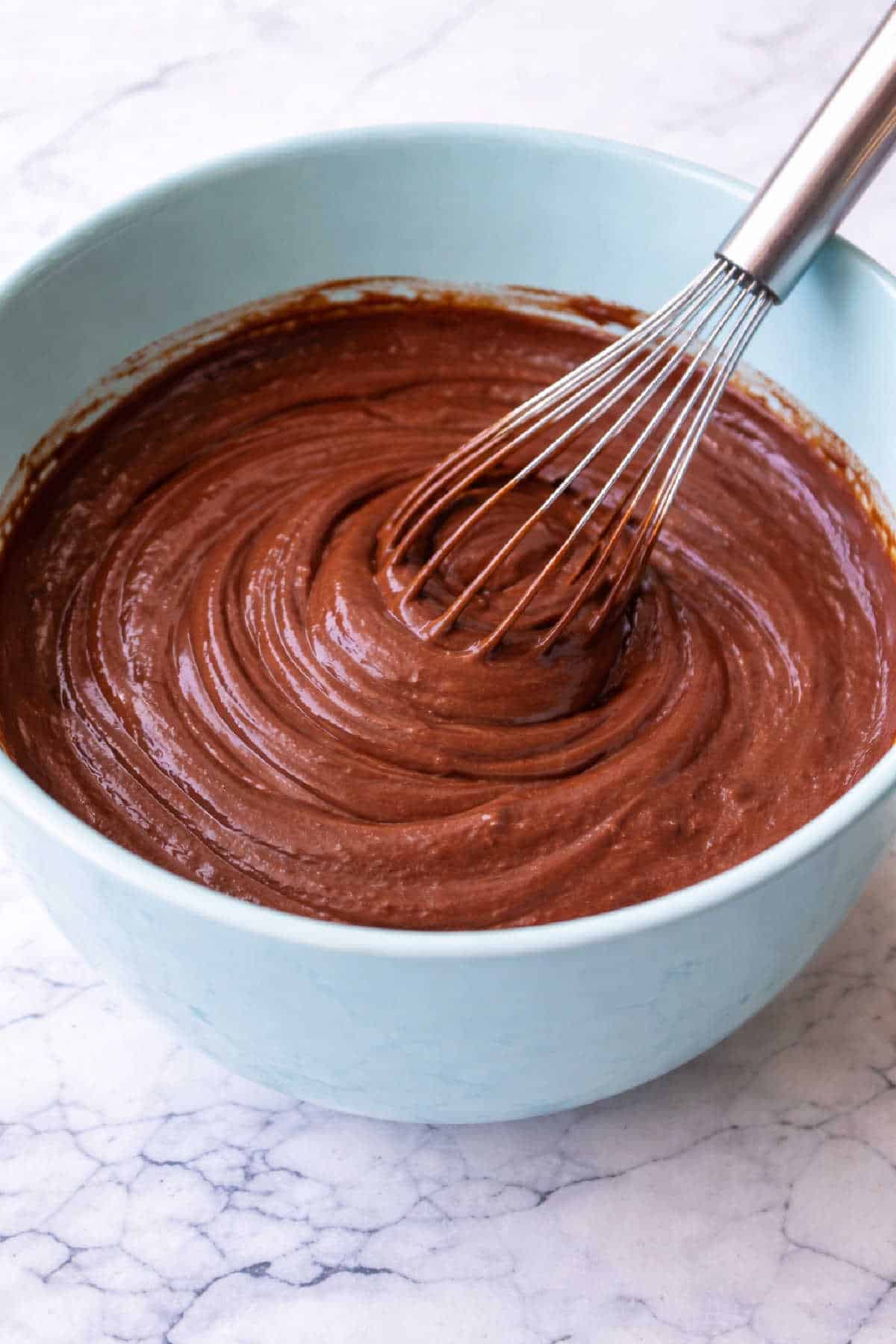 A metal whisk mixing smooth chocolate pudding in a light blue bowl on a marble countertop.