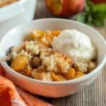 A bowl of peach cobbler topped with a scoop of vanilla ice cream, served with a spoon on a wooden table.