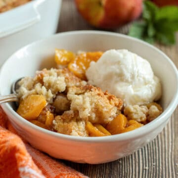 A bowl of peach cobbler topped with a scoop of vanilla ice cream, served with a spoon on a wooden table.