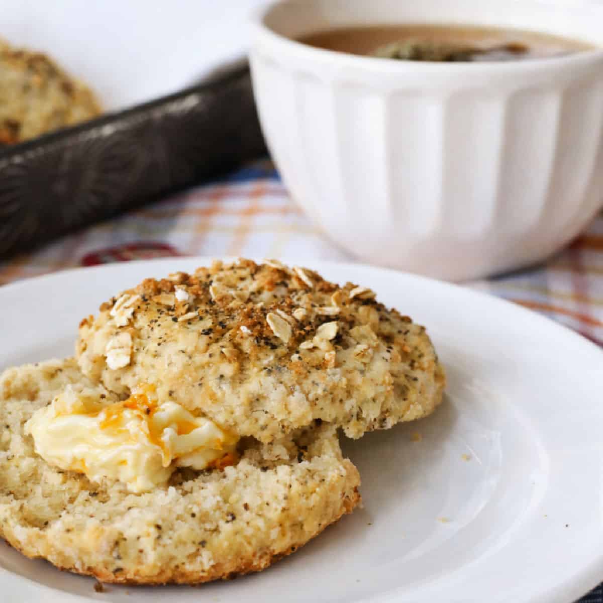 A biscuit split open with butter melting inside sits on a white plate, with a bowl of soup and another biscuit in the background.