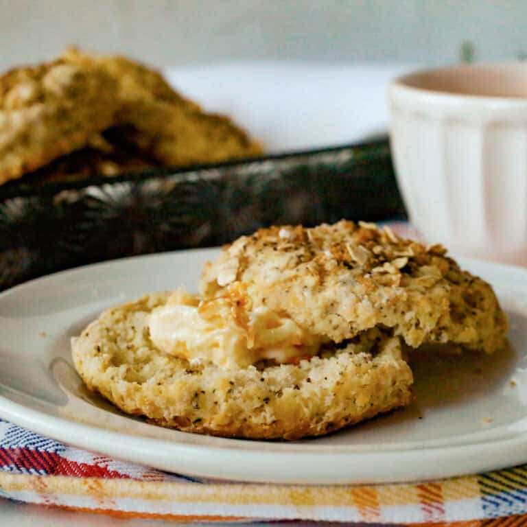 A split biscuit topped with butter and honey sits on a white plate. A tray of more biscuits and a light-colored cup are in the background.