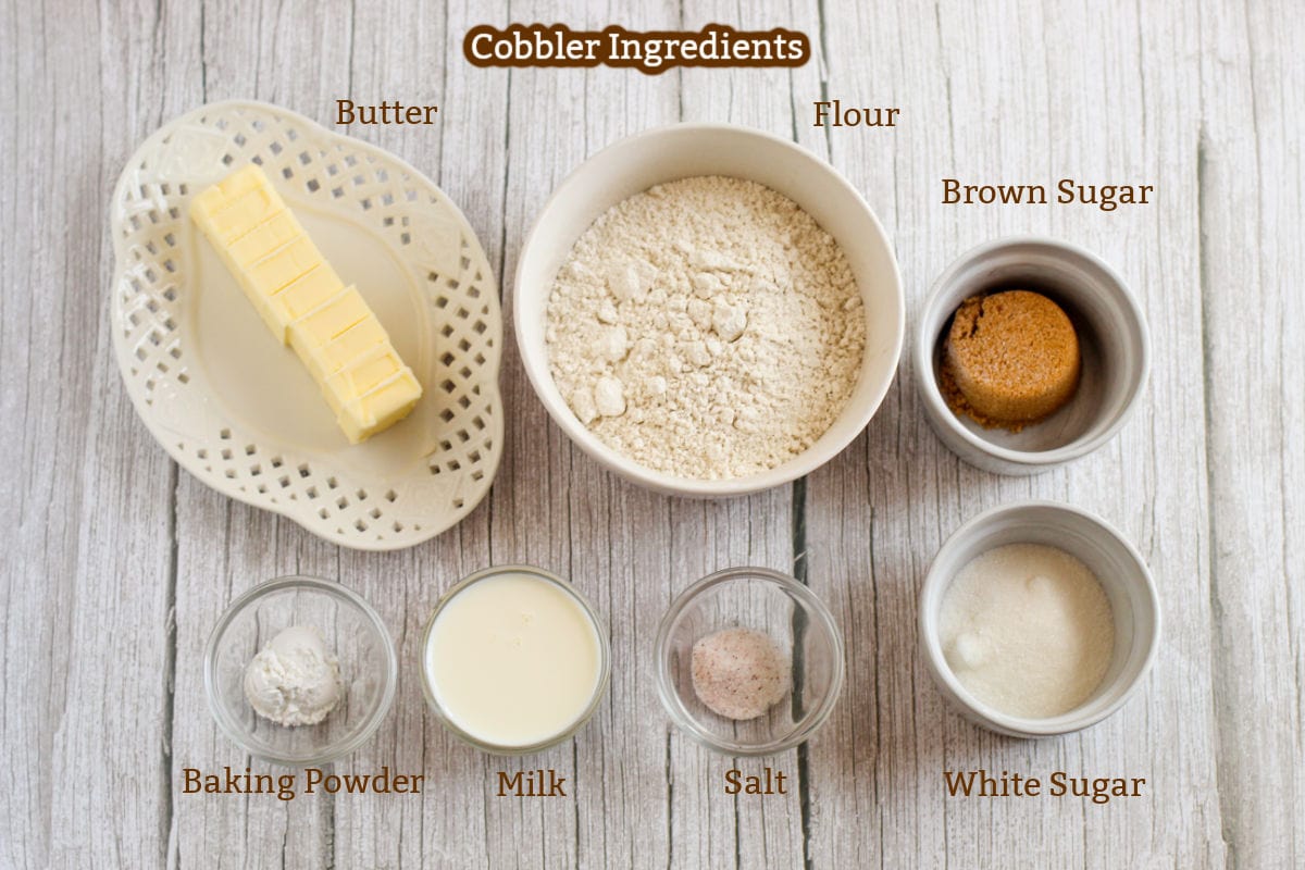Assorted cobbler ingredients on a wooden surface: butter, flour, brown sugar, baking powder, milk, salt, and white sugar, each in labeled bowls or plates.