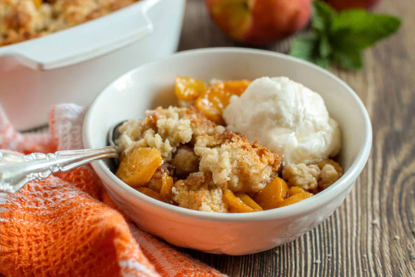 A bowl of peach cobbler with a scoop of vanilla ice cream and a spoon, placed on a wooden table next to an orange cloth.