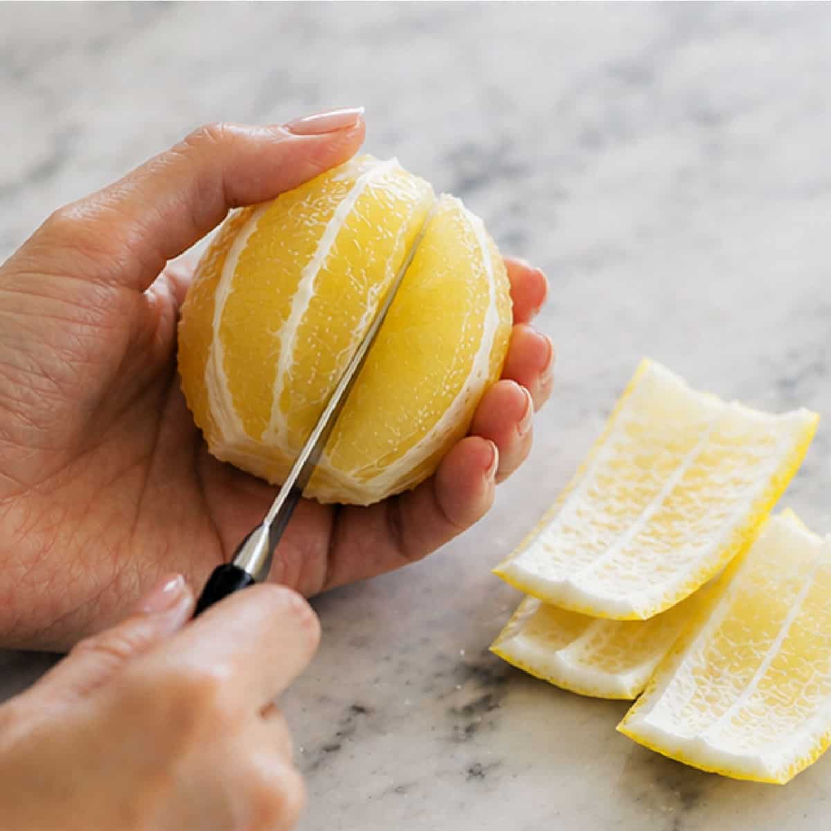 A person slices a peeled lemon with a knife on a marble surface, with lemon peels set aside.