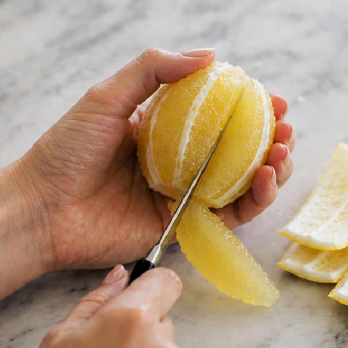 A person uses a knife to segment a peeled grapefruit, holding the fruit in one hand and cutting along the membrane with the other.