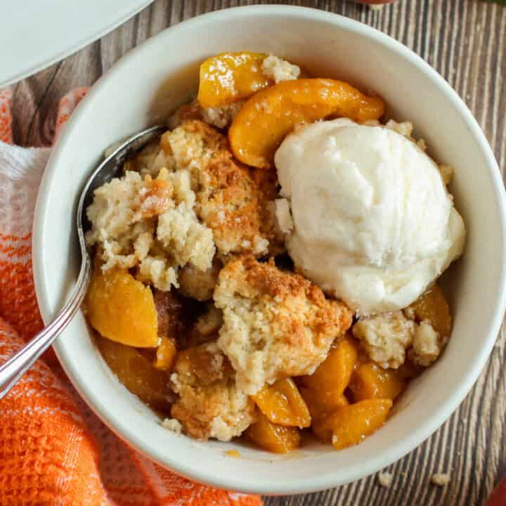 A bowl of peach cobbler topped with a scoop of vanilla ice cream, served with a spoon on a wooden table.