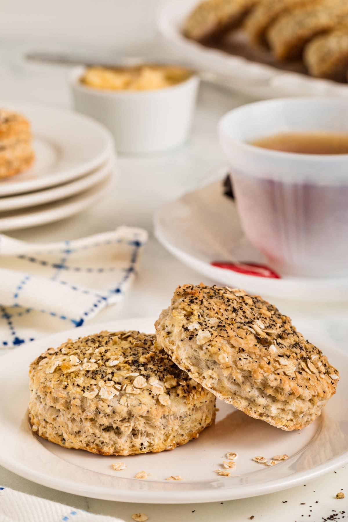 Two oat biscuits topped with seeds on a white plate, with a cup of tea, napkin, and other biscuits visible in the background.