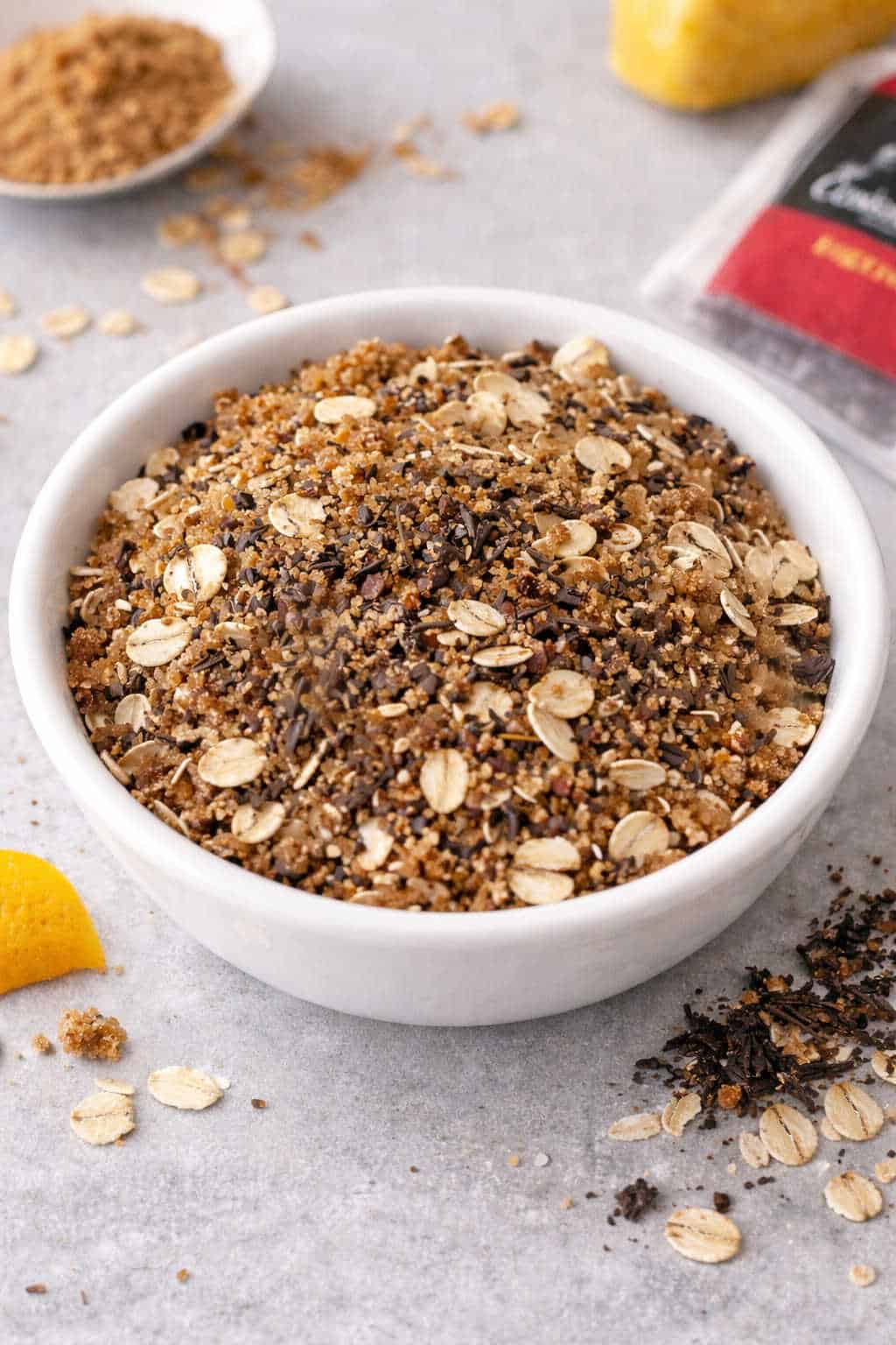 A white bowl filled with a mixture of brown sugar, oats, and spices sits on a light surface, with some oats and crumbs scattered nearby.
